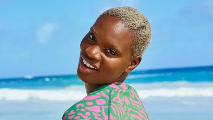 Ashley Byrd poses in a green and pink one-piece in front of the vibrant blue ocean.